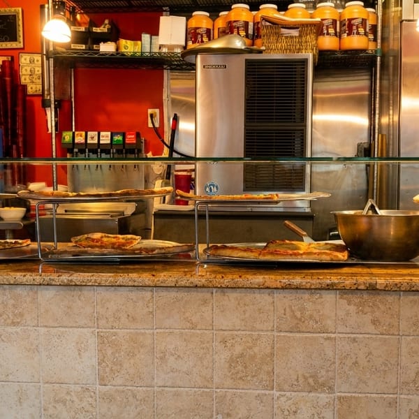 Pizza slices and whole pizzas displayed on a counter with a red wall and stainless steel appliances.