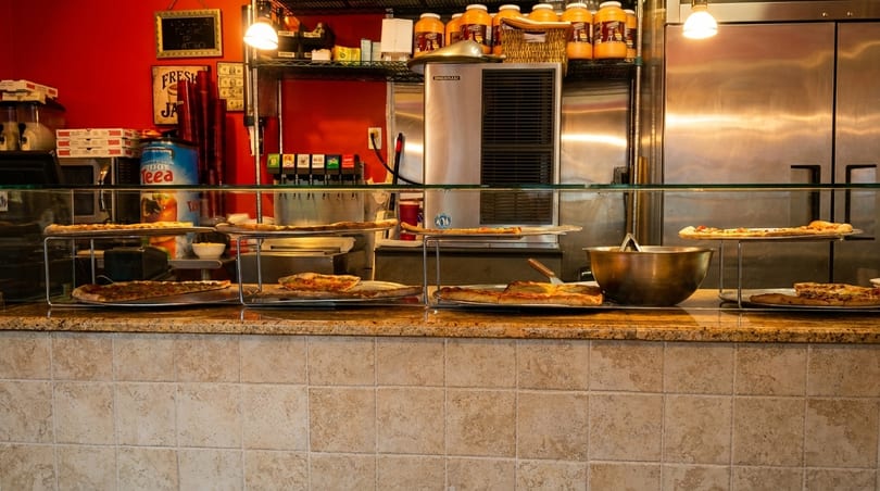 Pizza slices and whole pizzas displayed on a counter with a red wall and stainless steel appliances.