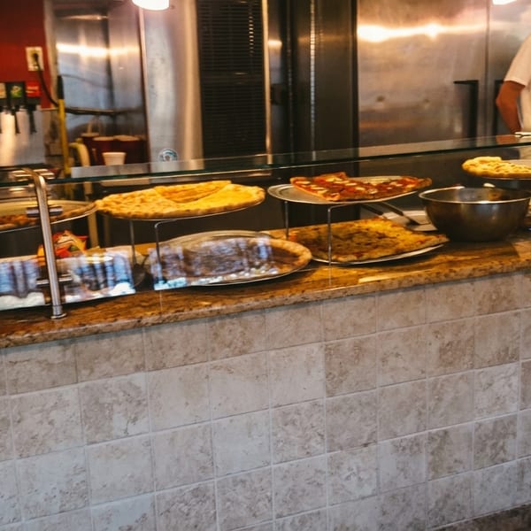 A pizza counter with various pizzas on display, and a person in the background.