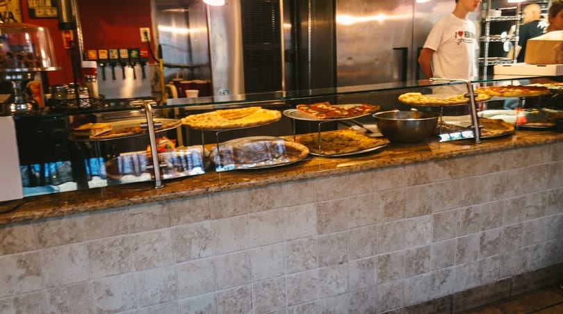 A pizza counter with various pizzas on display, and a person in the background.