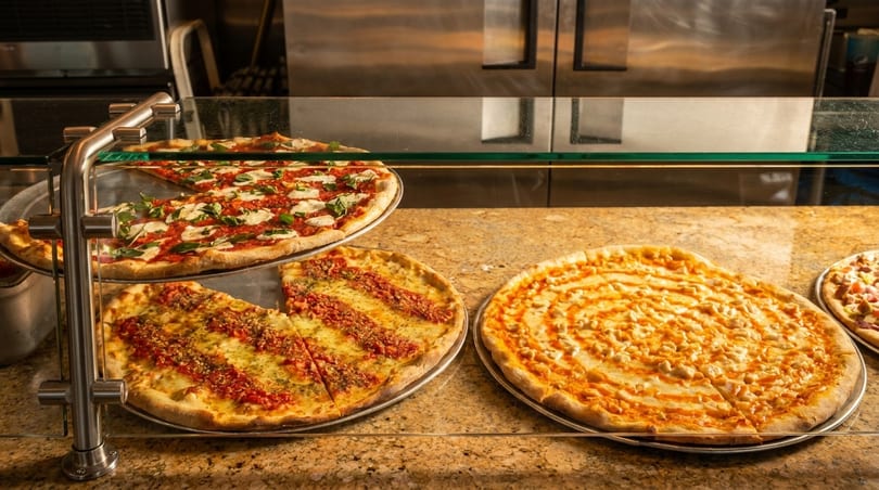 Three large pizzas, including Margherita and Sicilian, displayed under a glass counter.