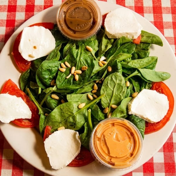 Spinach salad with tomatoes, mozzarella, pine nuts, and two dressing cups on a red and white checkered tablecloth.