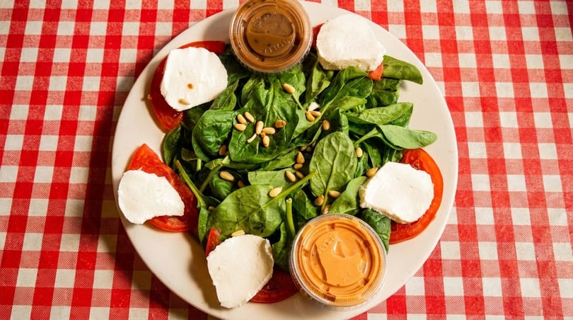 Spinach salad with tomatoes, mozzarella, pine nuts, and two dressing cups on a red and white checkered tablecloth.